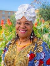 Woman, smiling, in blue and gold patterned top, and with white headpiece, standing in front of plants and building.