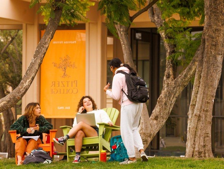 Standing student with backpack talks to two seated students on the Mounds with Pitzer banner in the background.